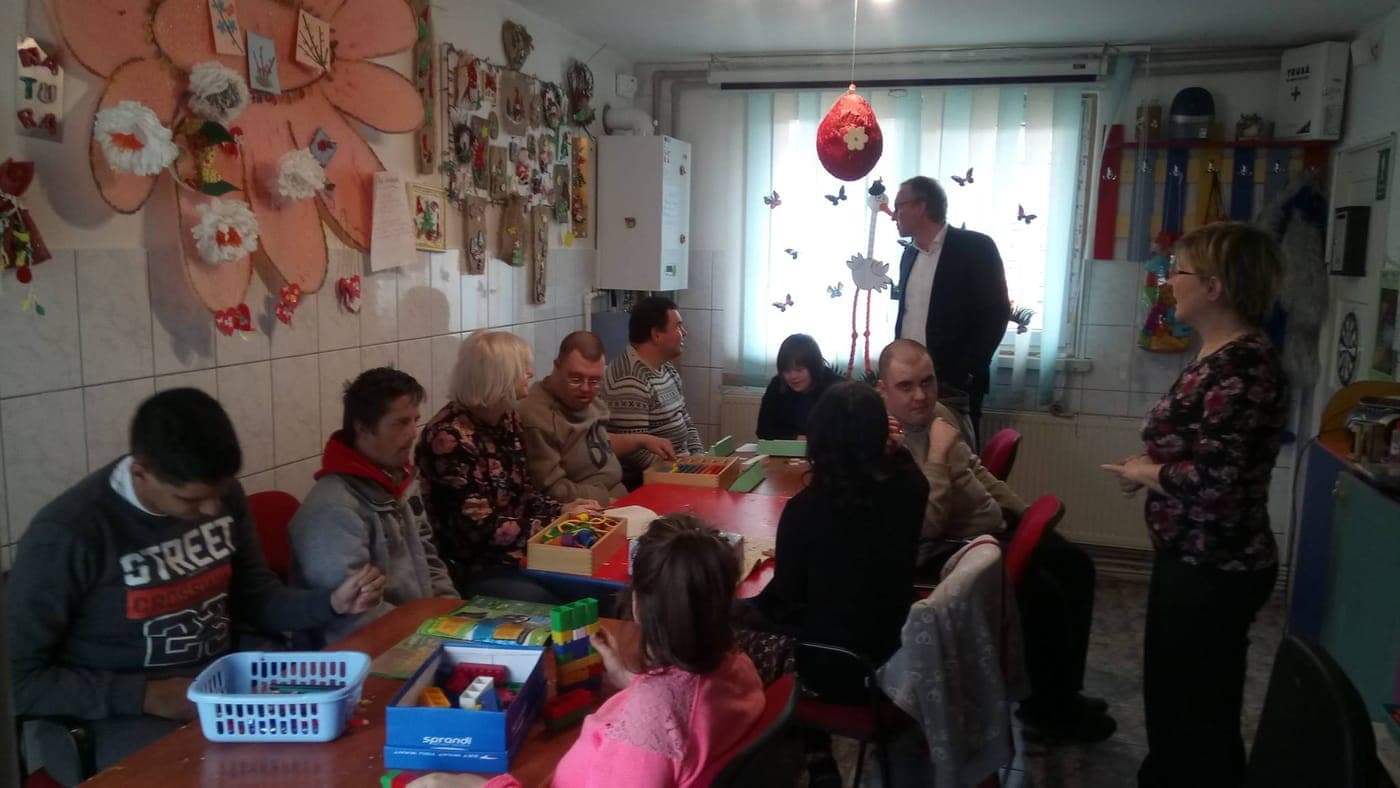 Persoane adulte participând la activități la mese, într-o sală de centru de zi decorată cu lucrări handmade.
