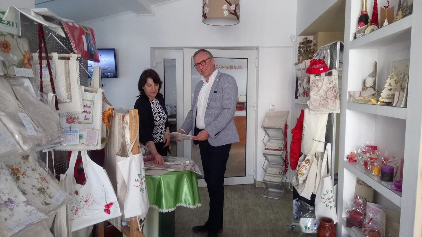 Două persoane examinând produse textile într-un atelier protejat, printre sacoșe și obiecte lucrate manual.