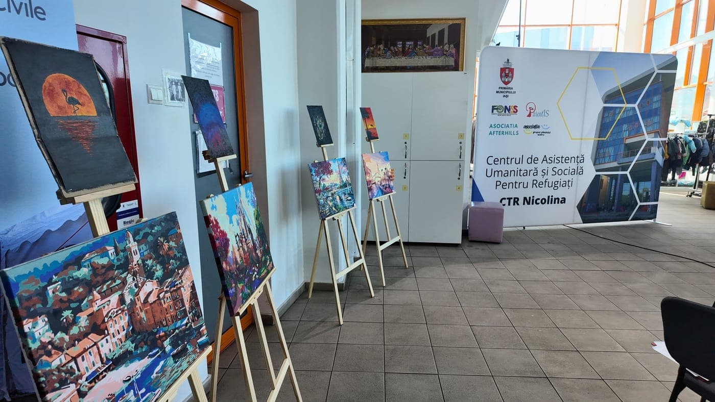 Expoziție de picturi pe șevalete, lângă un panou al Centrului de Asistență Umanitară și Socială pentru Refugiați CTR Nicolina.