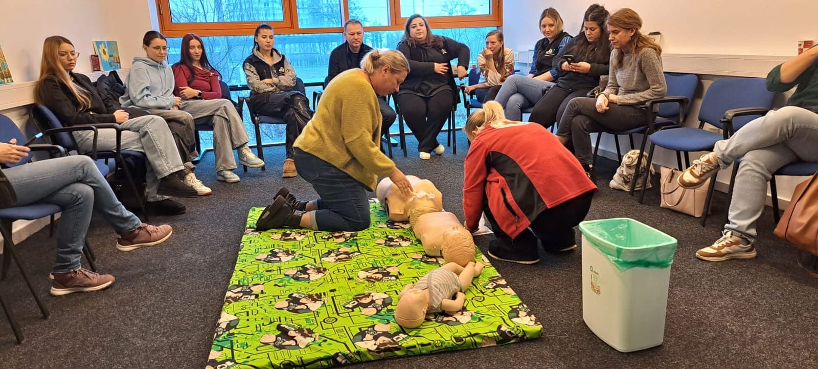 Instructor demonstrând manevre de prim ajutor pe un manechin, în fața unui grup așezat în semicerc.
